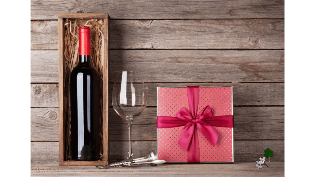 a bobble ot red wine in a wooden box next to a wine glass and a wrapped gift next to it